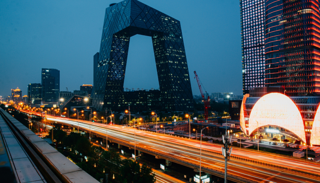 Cityscape at dusk featuring modern skyscrapers, including a uniquely shaped building, with light trails from traffic on a busy road.